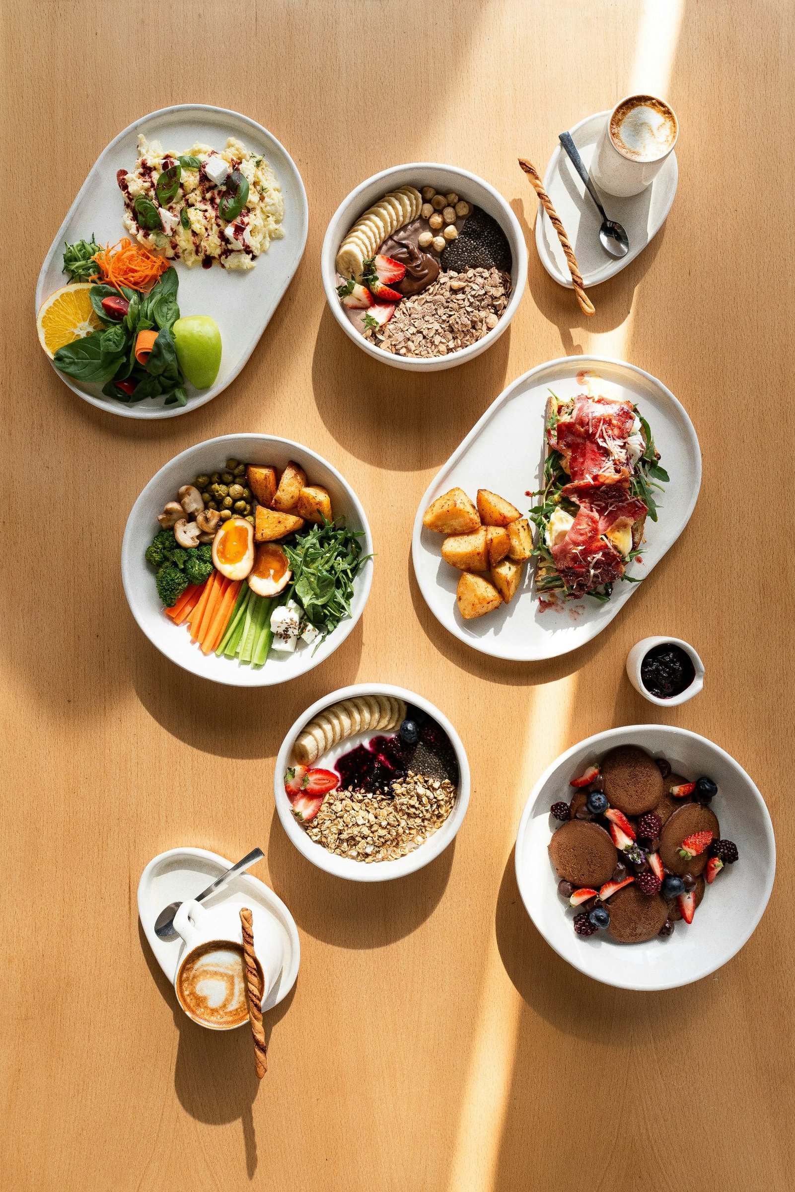 Overhead view of colorful healthy breakfast bowls, plates and coffee on a sunlit wooden table.